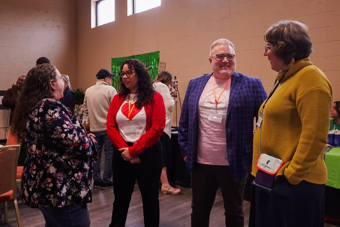 Four people in conversation at a social event, standing indoors with a tan brick wall background and some tables in the room.
