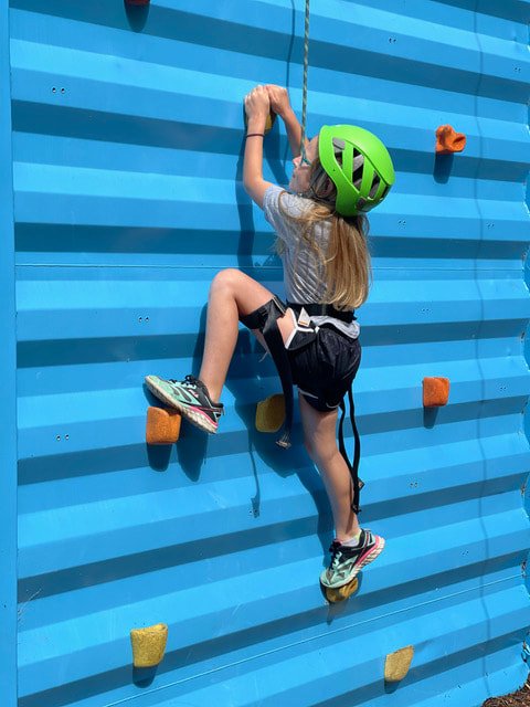Young blonde girl climbing blue climbing wall wearing bright green helmet and climbing harness