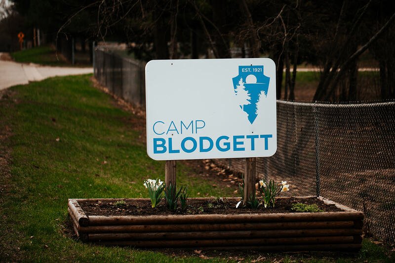 Sign for Camp Blodgett with a logo of trees and sun, in a flower bed surrounded by wooden border, with grass and a fence in the background.