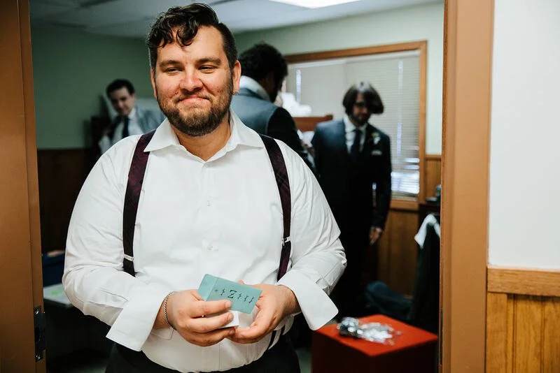 A man in a white shirt with suspenders holding tickets or cards, smiling, with three men in suits in the background in a room with wooden paneling and blinds.