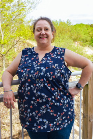 A woman smiling outdoors, standing on a wooden bridge with green trees and shrubs in the background.