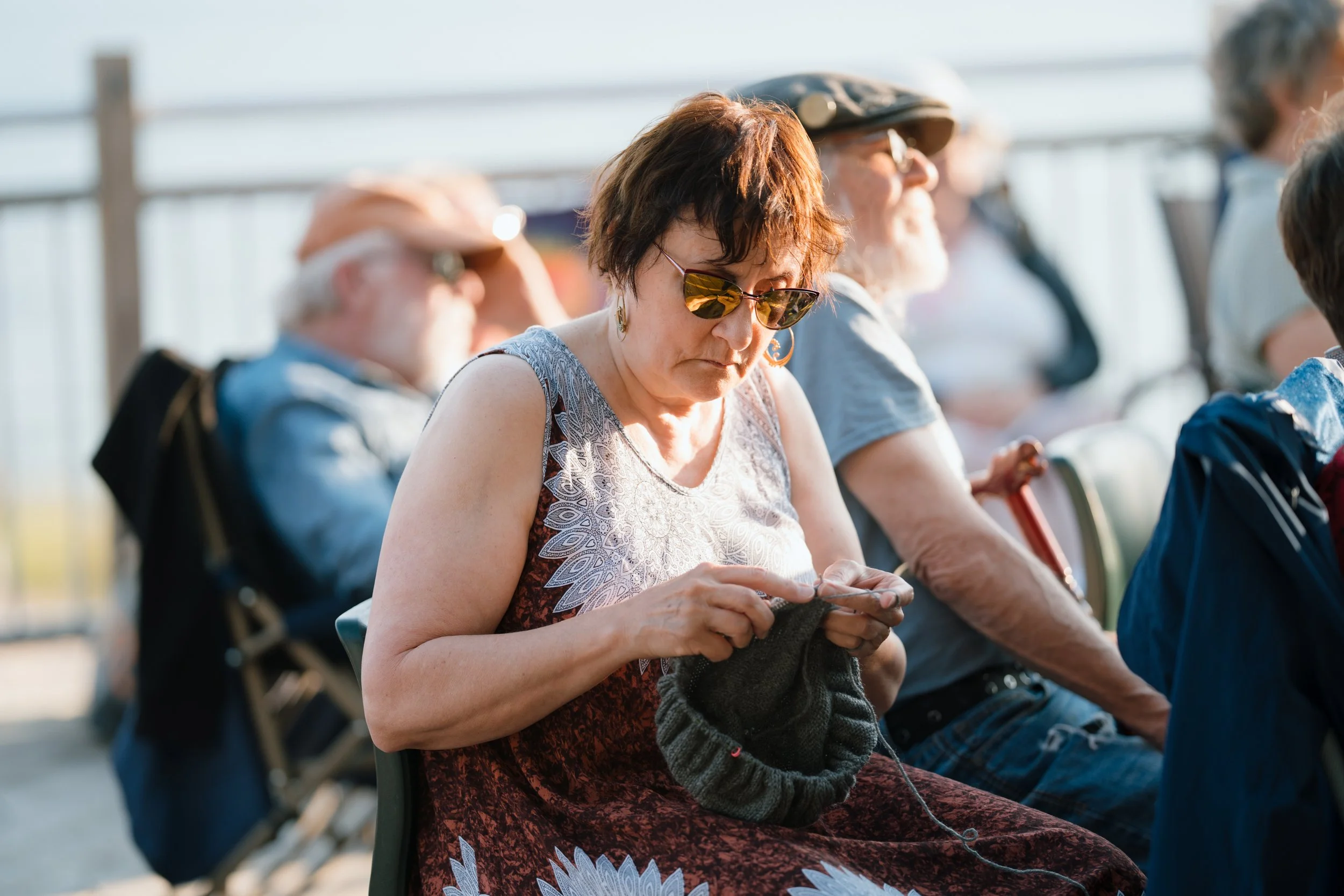 A woman with short brown hair, wearing sunglasses and a sleeveless dress, is knitting or sewing with yarn in her hands. She is sitting outdoors among other people, some wearing hats, on a sunny day near a metal or wooden fence.