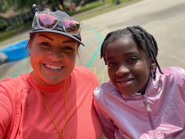 A woman and a girl are smiling for a selfie outdoors. The woman is wearing a pink shirt, sunglasses on her head, and a hat. The girl is wearing a shiny pink jacket and has braided hair. The background shows a park or playground area.