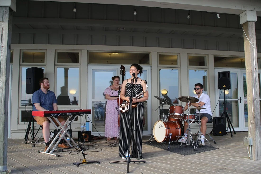A band performing on a wooden deck, with a female singer in striped overalls singing into a microphone, a keyboard player, a drummer, and a backup vocalist, with large windows and a door behind them.