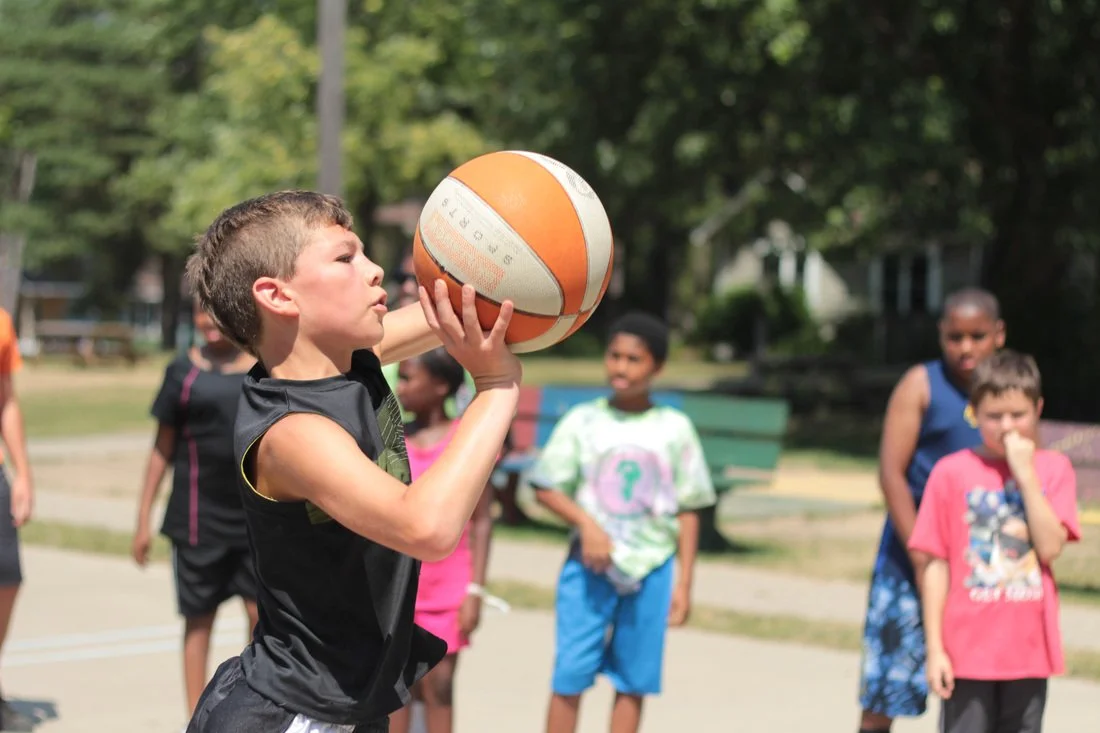 A young boy in a black sports jersey shooting a basketball at an outdoor court with other children watching.