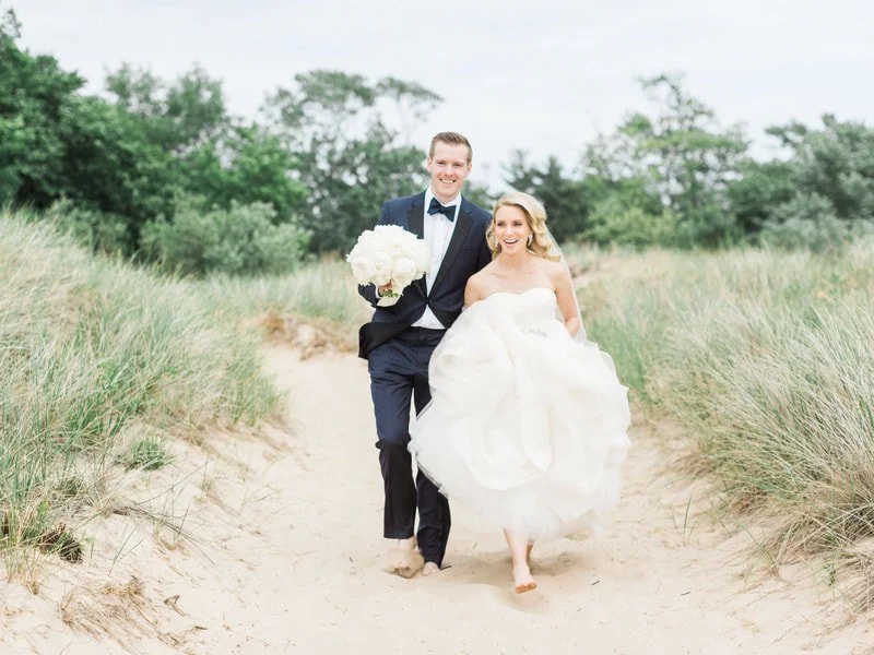 Bride and groom barefoot walking on sandy path through grassy dunes, bride holding her gown, groom carrying a bouquet, smiling, outdoors on a cloudy day.