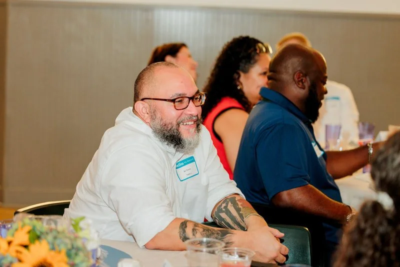 Group of people seated at a table, smiling and engaging in an event or meeting, with flowers and beverages on the table.