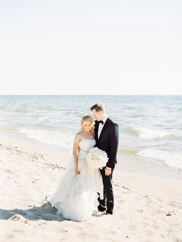 A bride and groom standing on the beach, with the ocean and blue sky in the background. The bride is wearing a white wedding dress and holding a bouquet of white flowers. The groom is dressed in a tuxedo. They are close together, looking affectionate