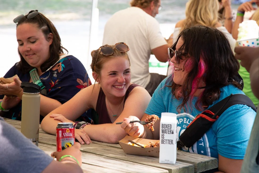 Group of women at a picnic table enjoying food and drinks during a gathering near the beach, smiling and chatting.