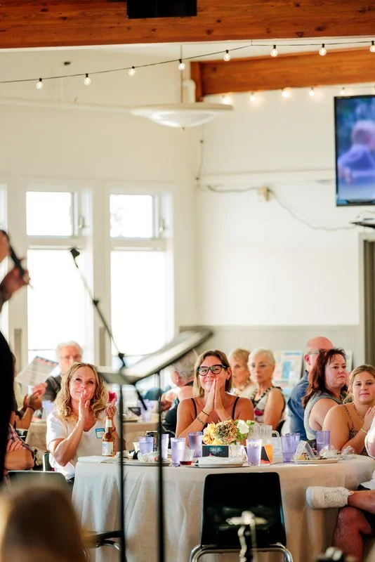 People sitting at round tables with floral centerpieces, applauding and smiling at a speaker in a well-lit room with string lights and large windows.