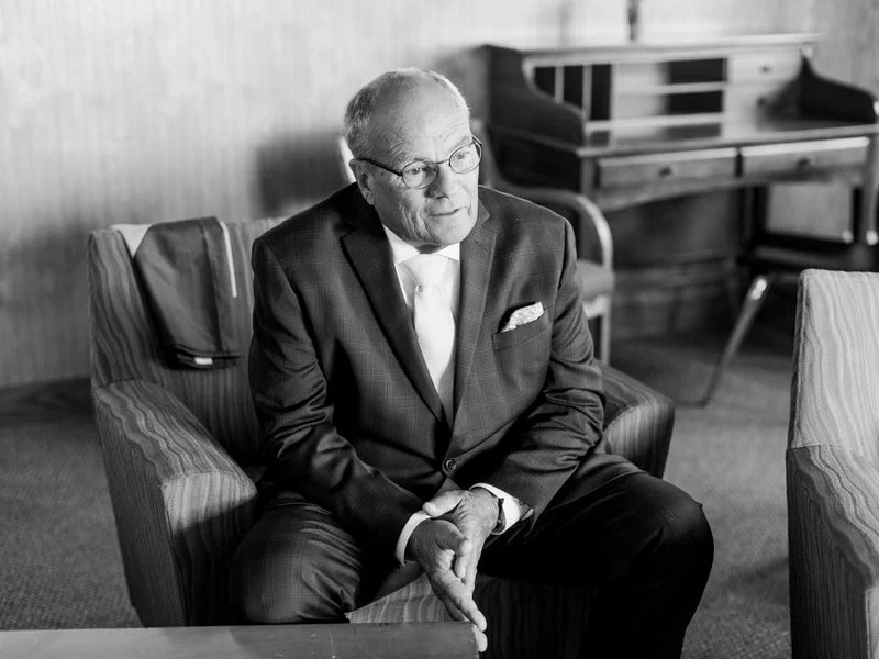 A black-and-white photo of an older man in a suit sitting on an armchair in an office or meeting room.