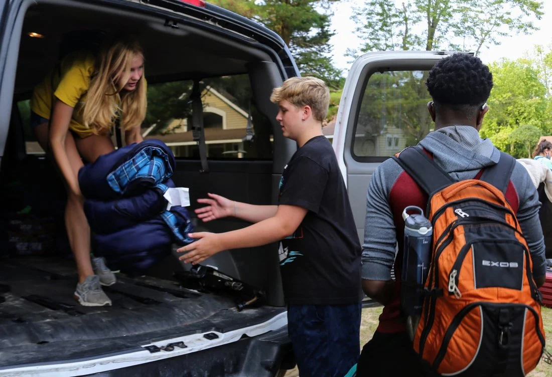 Three teenagers loading camping gear into the back of a vehicle outdoors surrounded by trees.