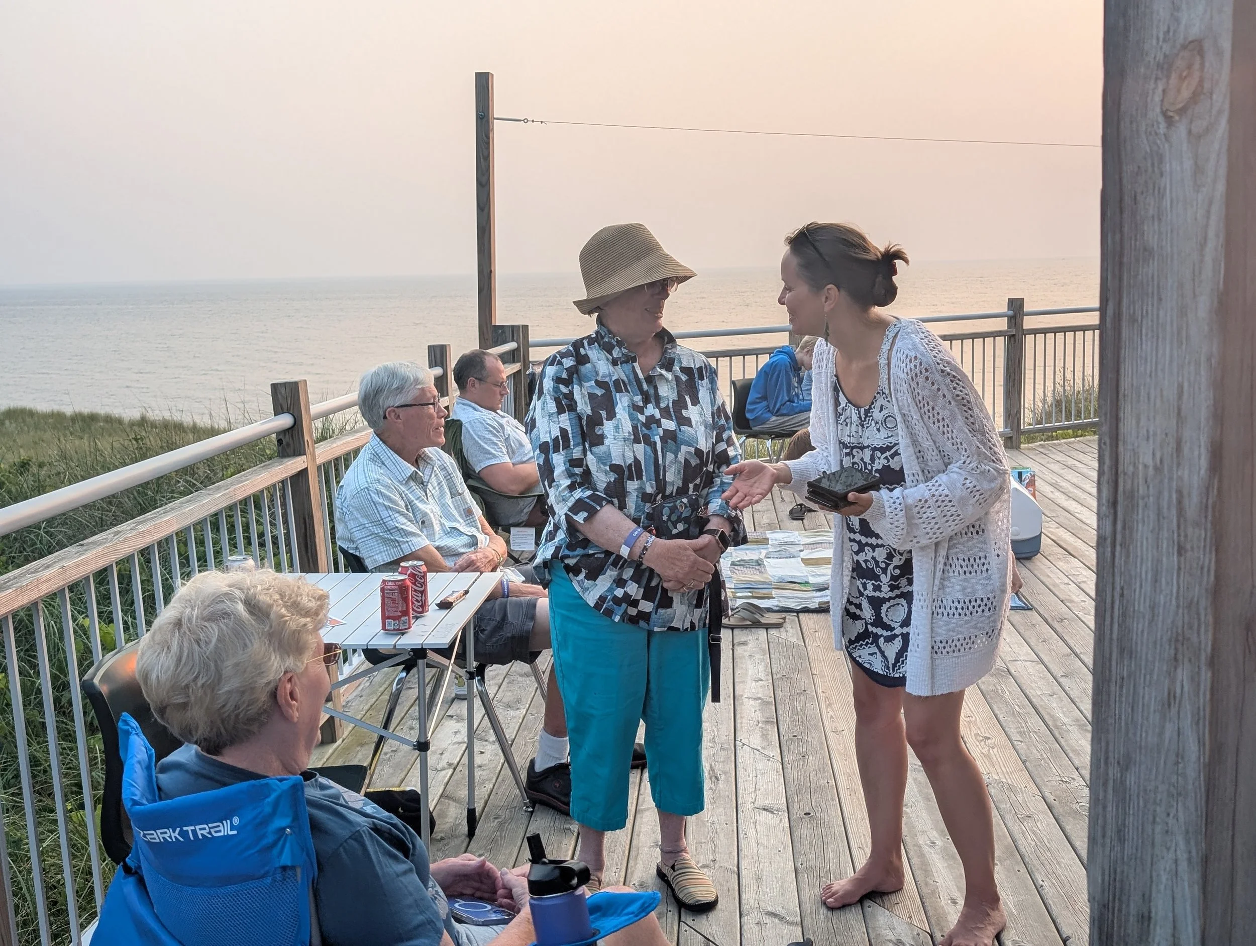 Group of people gathered outdoors on a wooden deck near the ocean at sunset, with some sitting and others standing, engaged in conversation, one woman holding a stack of photographs.