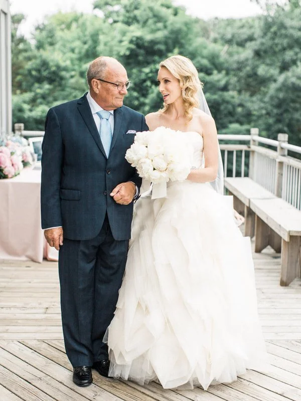 Bride in a white wedding dress holding a bouquet walking with an elderly man on a wooden deck outdoors during a wedding ceremony.