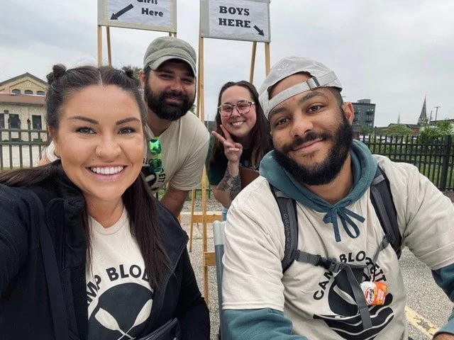 Four smiling people taking a selfie outdoors near a sign that says 'GIRLS HERE' and 'BOYS HERE' in front of a fence.