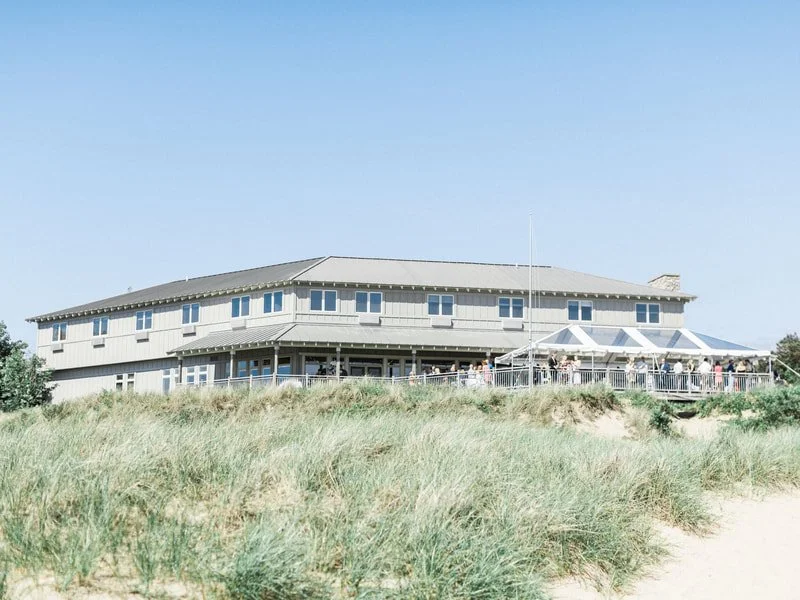 A large, two-story beachside building with a deck and outdoor seating area, surrounded by grassy dunes and located near a sandy beach.