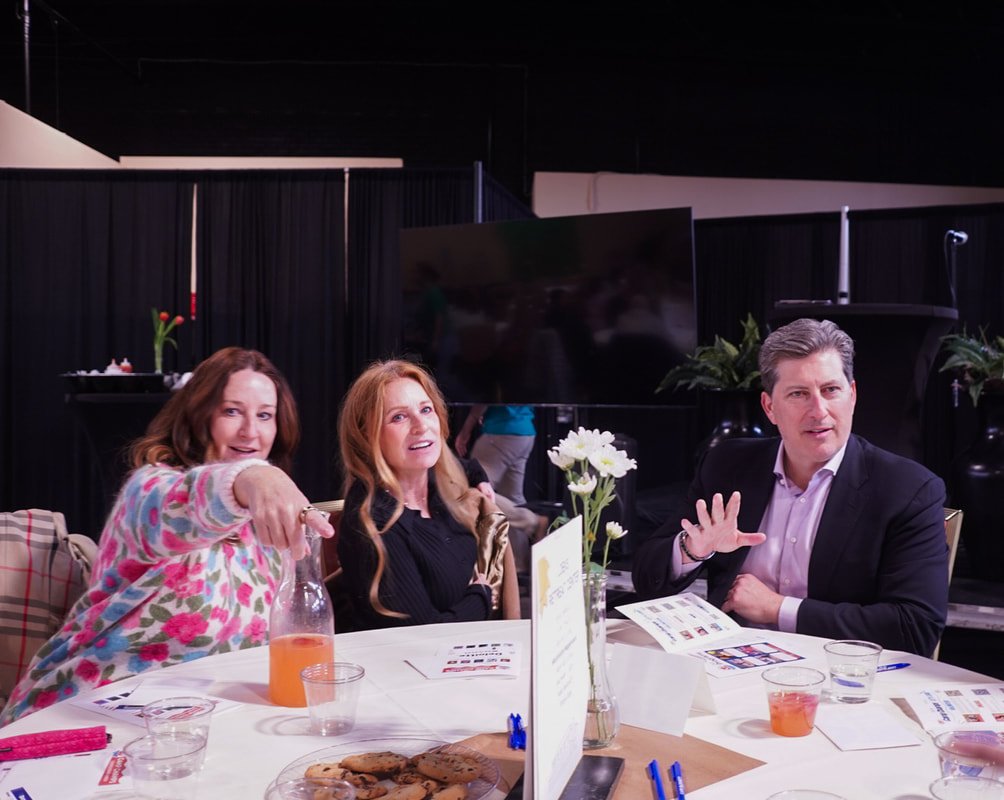 Three people sitting at a round table during a meeting or event, with one woman pointing towards the camera, a red-haired woman sitting in the middle, and a man on the right. There are flowers, drinks, talking points, and papers on the table.