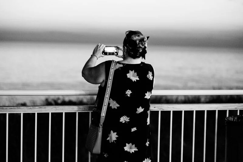 A woman taking a photo with her phone on a ferry or boat, overlooking the water, wearing a dress with large floral prints and a purse with a Michael Kors strap.