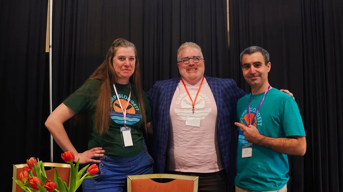 Three people posing for a photo at an indoor event, standing in front of a black curtain. The person on the left is a woman with long brown hair, wearing a green Camp Blogger t-shirt and a white lanyard. The person in the middle is a man with glasses