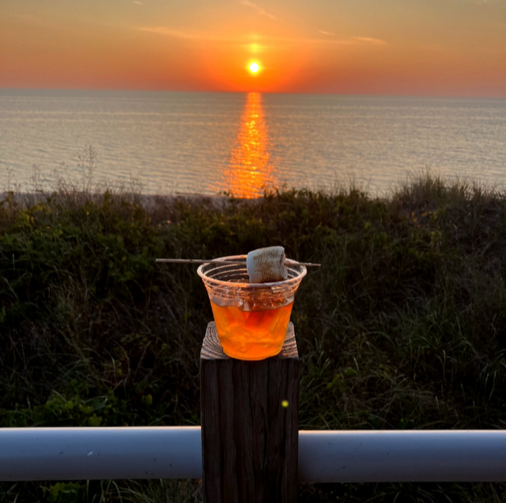 A sunset over the ocean with a drink and marshmallow on skewers in the foreground.