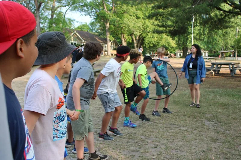 Group of children participating in a game of jump rope outdoors at a camp or park, with an adult supervising.