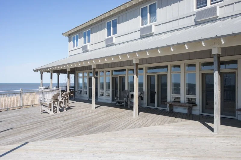 Beachside wooden deck with benches and chairs, overlooking the ocean, attached to a white coastal house with large windows and a balcony.