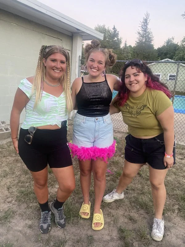 Three young women standing outdoors near a chain-link fence, smiling at the camera, dressed casually with playful accessories and outfits, in a backyard with trees and a pool in the background.