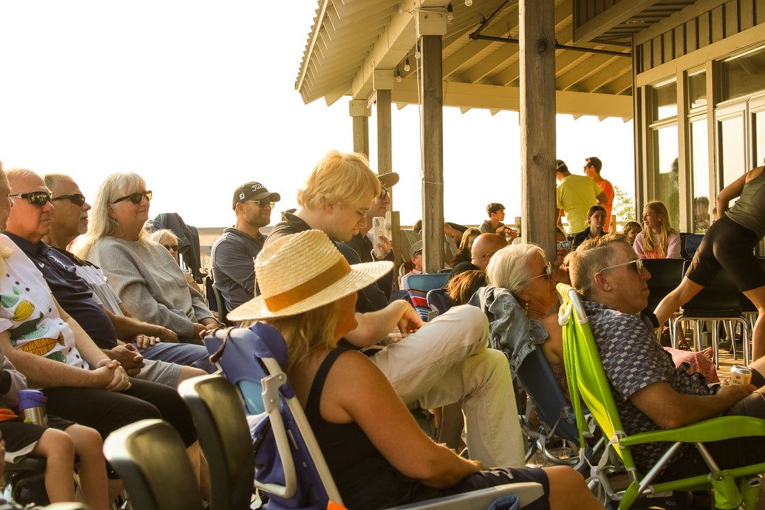 Group of people sitting on chairs on an outdoor patio, some wearing sunglasses and hats, facing a building with large windows. The scene appears to be during a sunny time of day.