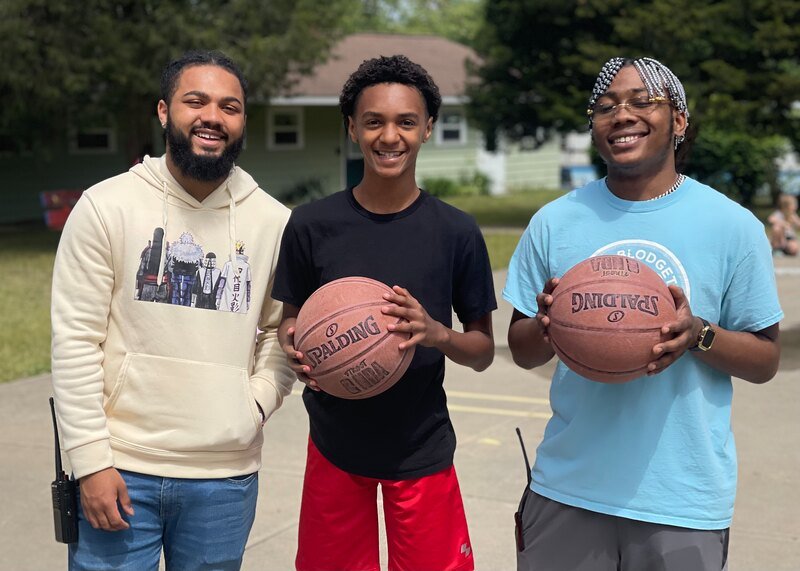 Three young men smiling outdoors, two holding basketballs, in a park or playground with trees and a small building in the background.