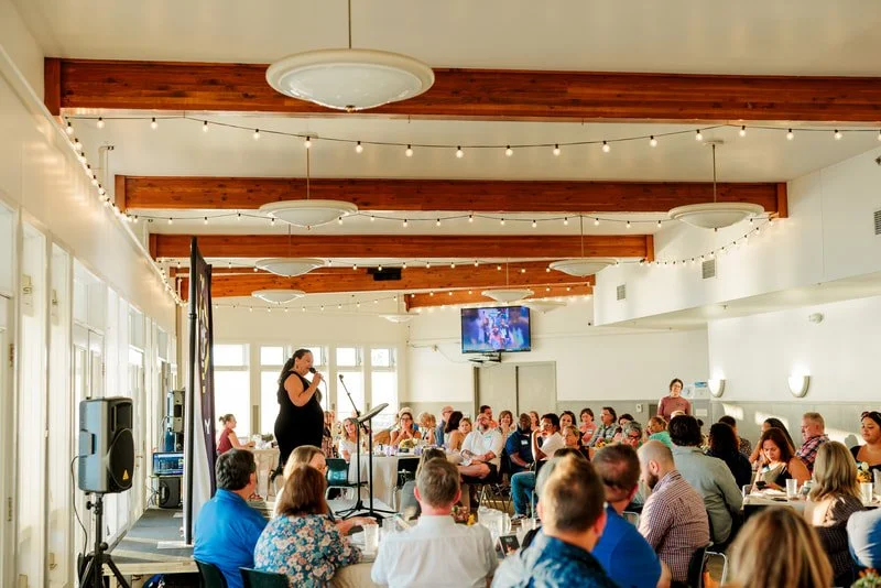 A woman is speaking at a microphone during a formal event or conference in a bright, modern room with wooden beams, string lights, and a TV monitor, while an audience of men and women sit at tables listening.