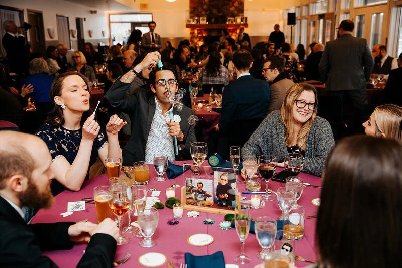 People celebrating at a formal event or party, sitting at a table with drinks, decorations, and photos, with a fireplace and Christmas tree in the background.