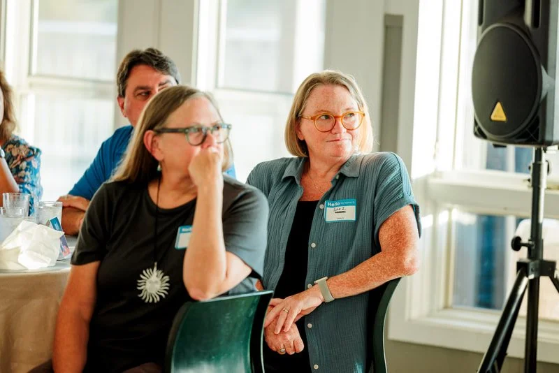 Group of people sitting at a table during a meeting or conference, with one woman wearing glasses and a name tag, and a man behind her, all in a well-lit room with large windows.