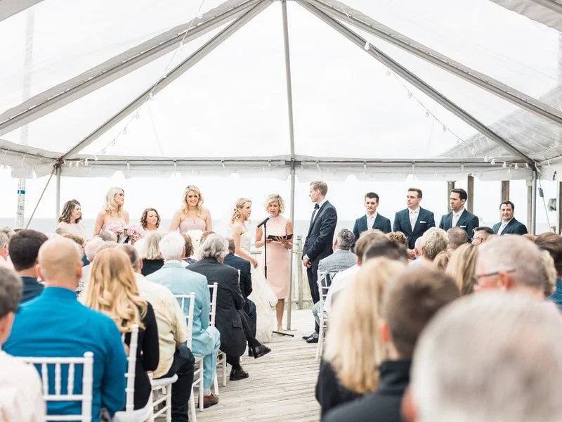 Wedding ceremony taking place under a tent, with a bride and groom standing at the altar, surrounded by bridesmaids and groomsmen, with seated guests observing.