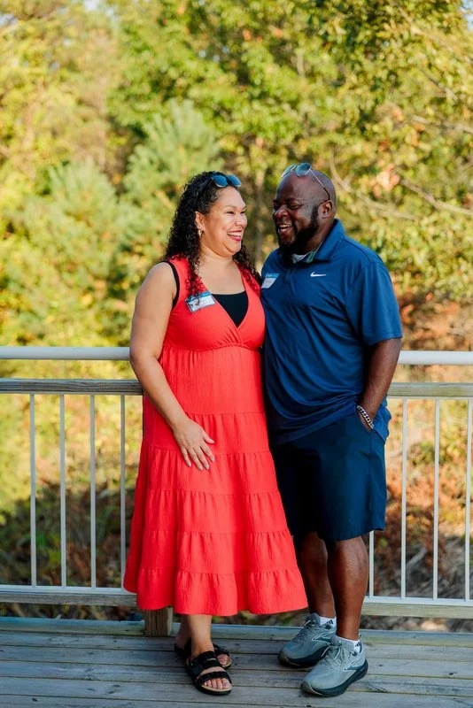 A woman in a red dress and a man in a blue shirt and black shorts, smiling and standing close together outdoors.