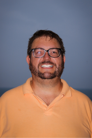 A man with glasses, a beard, and short dark hair smiling, wearing a peach-colored polo shirt, standing against a blue background.