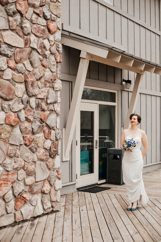 A bride in a white wedding dress holding a bouquet standing outside a building with gray wooden siding and a stone wall, in front of a glass door.