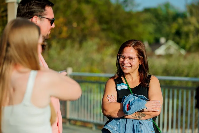 Smiling woman with glasses and a sleeveless top standing outdoors with arms crossed, holding a denim jacket and a drink, talking to two people in a sunny park setting.