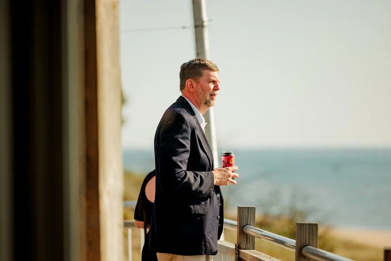 Man in a dark blazer holding a colorful soda can, standing by a wooden railing near the beach, with the ocean in the background.