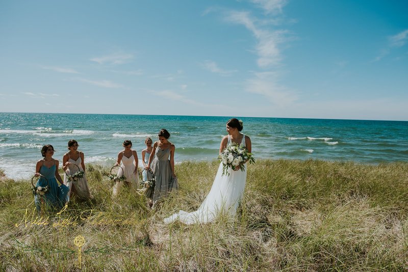 A bride in a white wedding dress holding a bouquet, standing on a grassy beach with five bridesmaids in dresses, near the ocean under a blue sky.