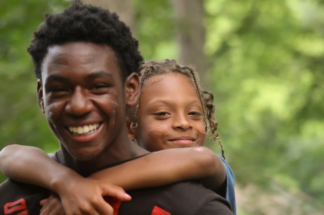 A young man is smiling and embracing a young girl in a park with green trees in the background.