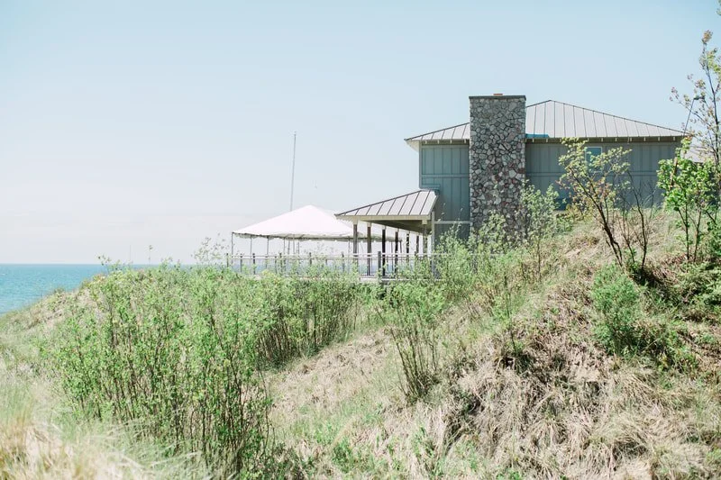 A house with a stone chimney on a grassy hillside near the ocean, with a patio and a white canopy.