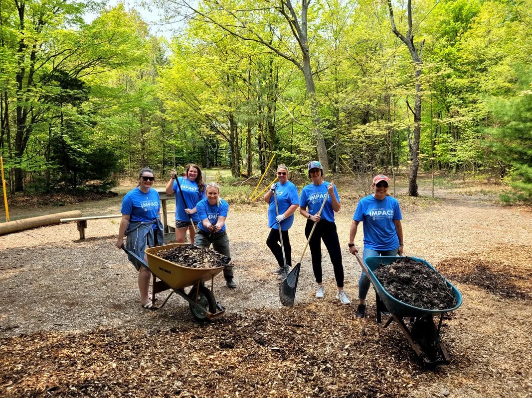 Group of people in blue t-shirts participating in a tree planting or park cleanup event in a wooded area, holding shovels and wheelbarrows filled with dirt.