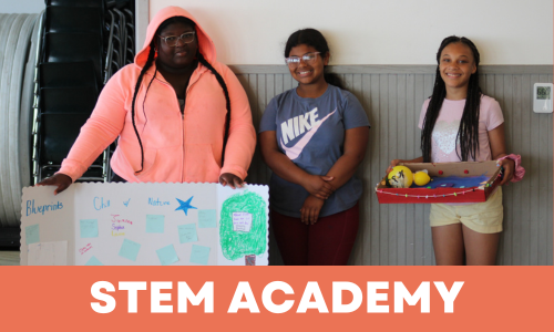 Three young girls standing inside a school hallway, holding a science project display and a tray of colorful objects. The display has writing and drawings, and the girls are smiling. The background includes a gray wall and a black chair.