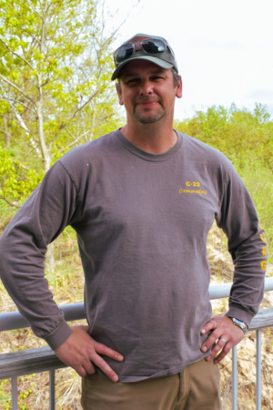 Man standing outdoors with a hand on his hip, wearing a gray long-sleeve shirt, sunglasses on his head, and a baseball cap, with trees and a cloudy sky in the background.