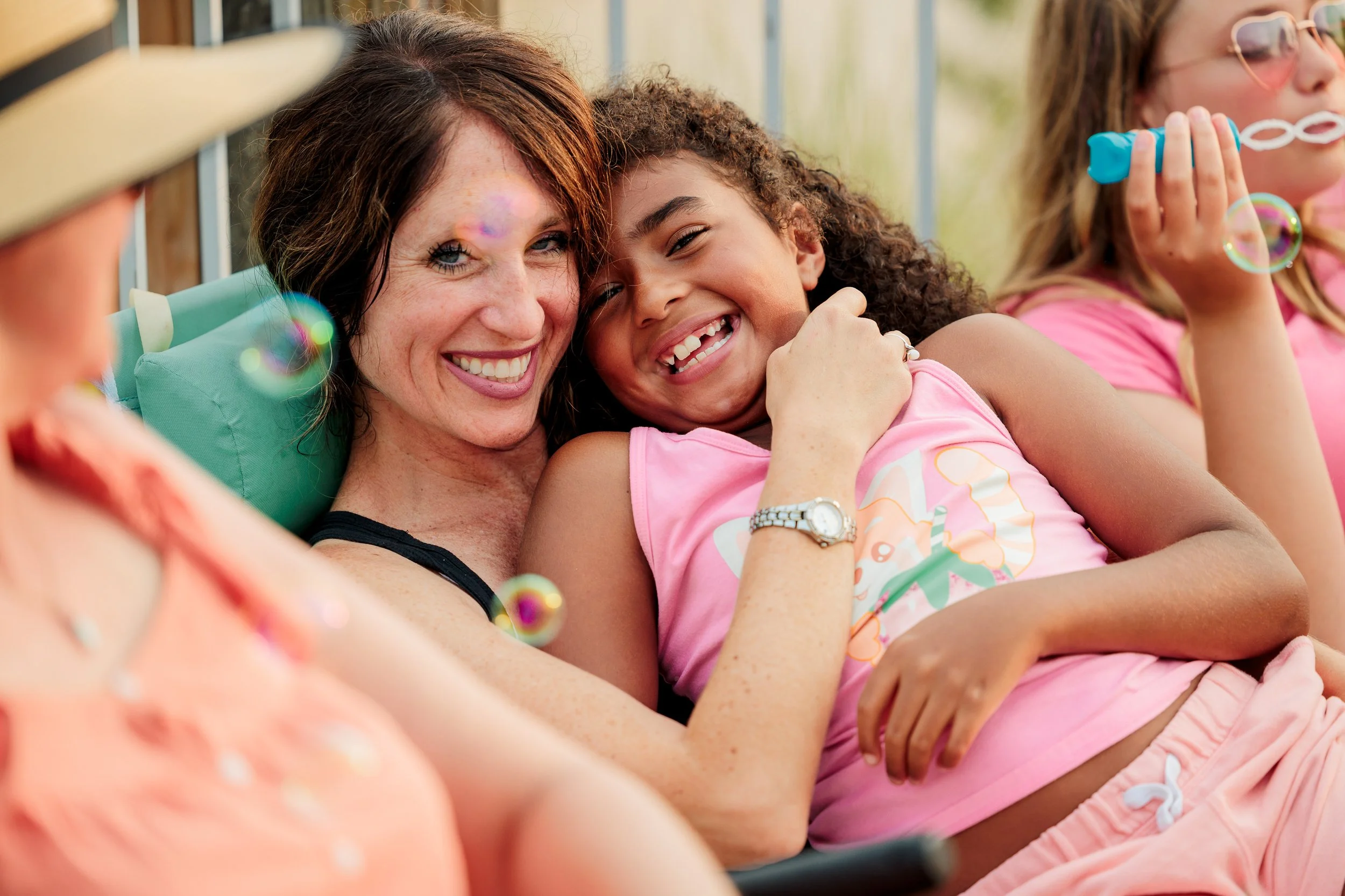 A group of smiling women enjoying a sunny day, with one woman and a young girl in pink shirts sharing a joyful moment. The girl is holding a bubble wand, and soap bubbles float around them.