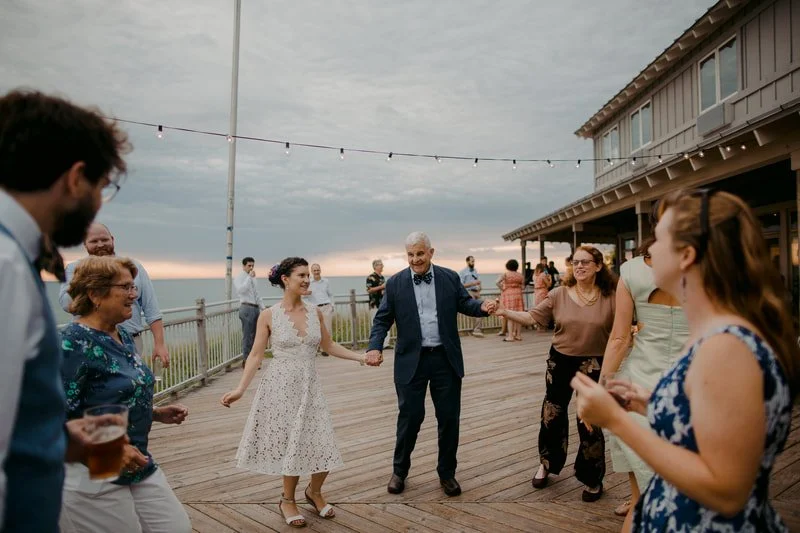People dancing and socializing on a wooden deck near the ocean, with a cloudy sky overhead, during a celebration or party, featuring string lights and a beachfront building.