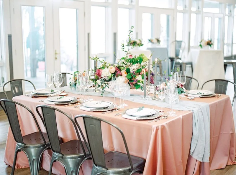 A decorated dining table set for a formal event with pink tablecloth, floral centerpiece, and place settings with plates, glasses, and silverware in a bright room with large windows.