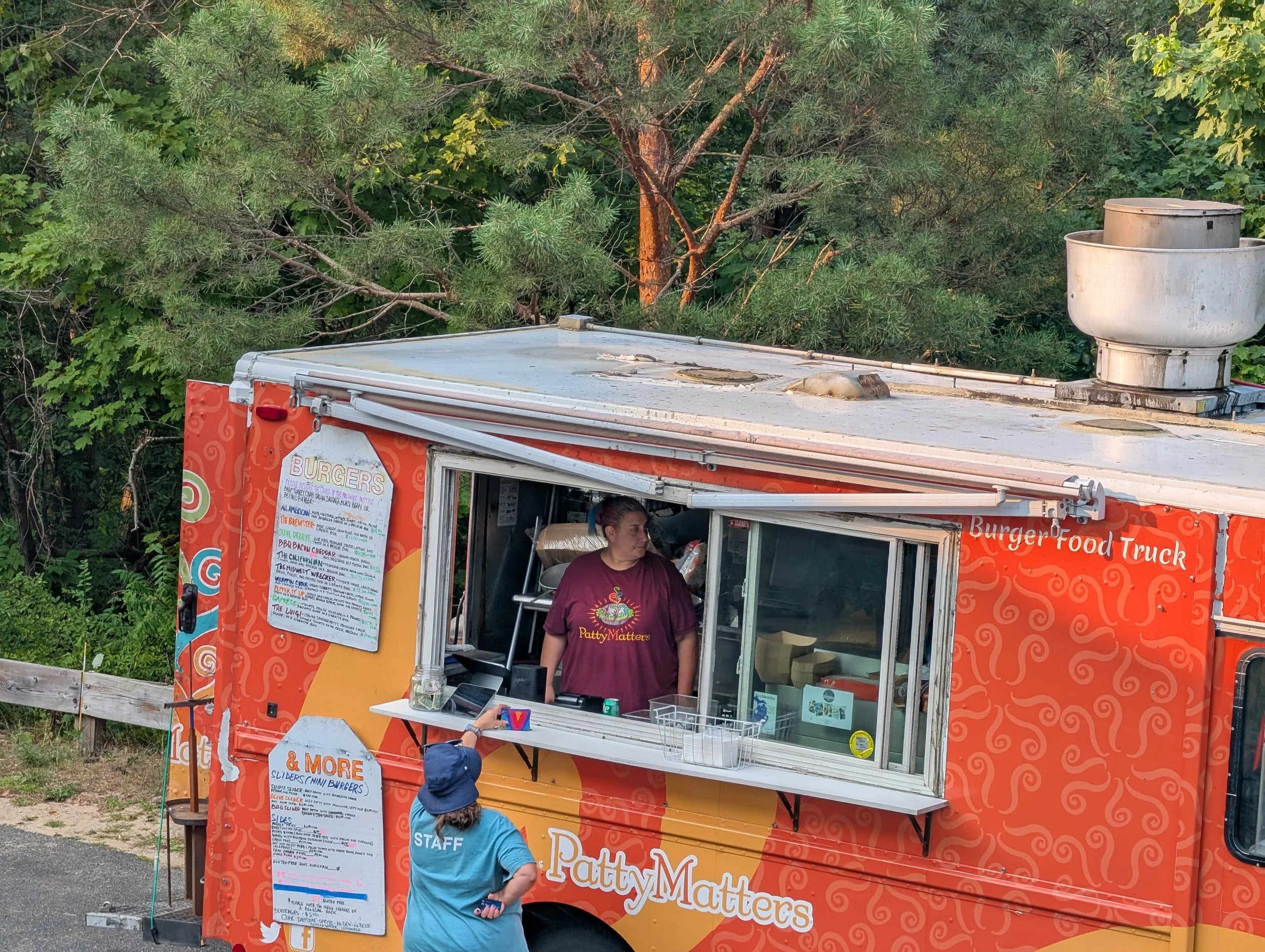A bright orange burger food truck named 'PattyMatters' is parked outdoors surrounded by green trees. A woman wearing a maroon shirt and purple gloves is working inside the truck. A customer wearing a blue shirt and a blue hat with 'STAFF' written on 