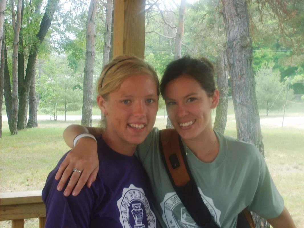 Two young women smiling, with the woman on the left wearing a purple Camp Blood shirt and the woman on the right wearing a green Camp Blood shirt and carrying a brown strap, standing outdoors with trees in the background.