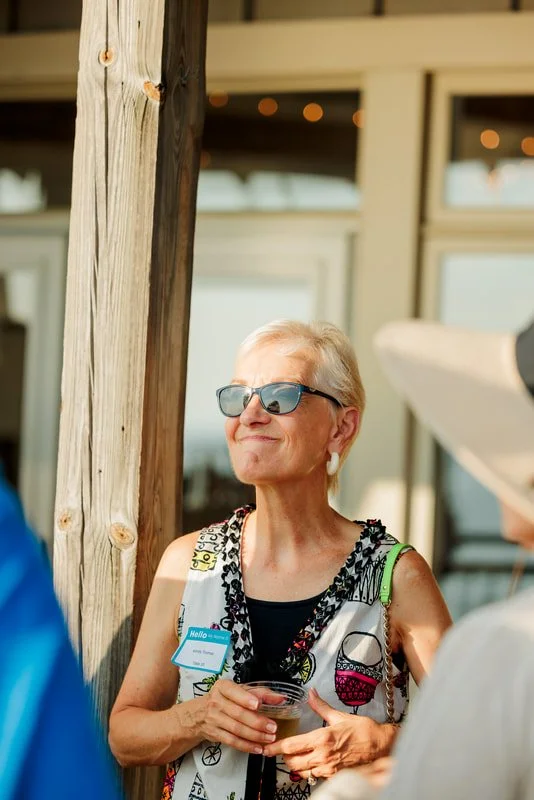 An elderly woman wearing sunglasses and a patterned sleeveless top, holding a drink, standing outdoors near a wooden post, with a building and string lights in the background.